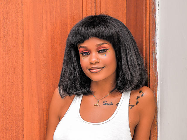 AlexaZane with dark, straight, shoulder-length hair, wearing a white tank top, has red eye makeup, a gold necklace, and a tattoo on her right shoulder. Her ass is not visible.