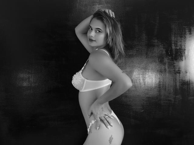 Fotografia in bianco e nero di Electraarding con seni di media grandezza, che indossa un reggiseno e uno slip di pizzo bianco, con una mano sulla testa, che guarda verso la fotocamera, mostrando un tatuaggio sull'anca destra, sfondo scuro..