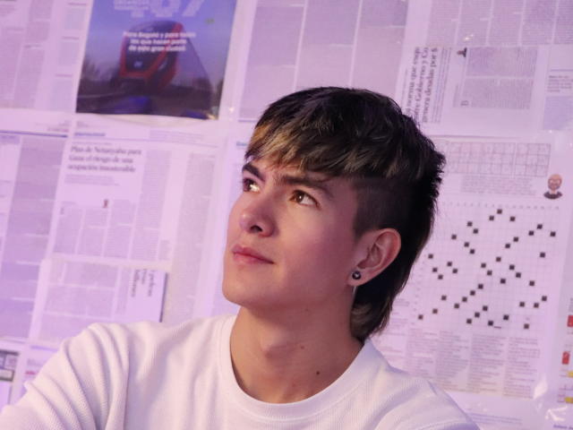 Archangel69, een jonge man met kort bruin haar, een wit shirt, oor piercings, die omhoog kijkt, met een lichte grijns, op een witte achtergrond met papieren. Zachte paarse verlichting, informeel, schattig..