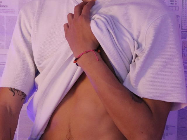 A camisa branca de Archangel69 está levantada, revelando um peito plano, com a pele morena, e uma pequena tatuagem no braço direito, além de uma pulseira rosa no pulso esquerdo..
