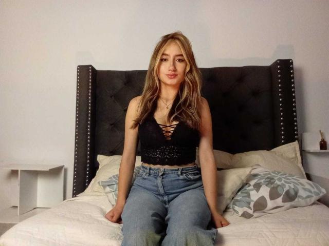SophiaPaz met blond haar, een zwarte top met veters en blauwe jeans, zittend op een bed met een donker hoofdeinde met studs, handen op het bed, kijkend naar de camera, met een lichte glimlach..