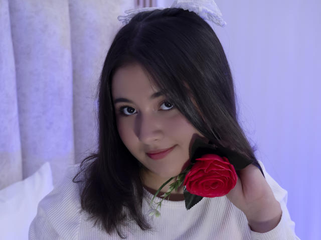 CatalinaRosel with dark hair, holding a red rose in front of her mouth, wearing a white lace headband and white ribbed shirt, smiling softly.