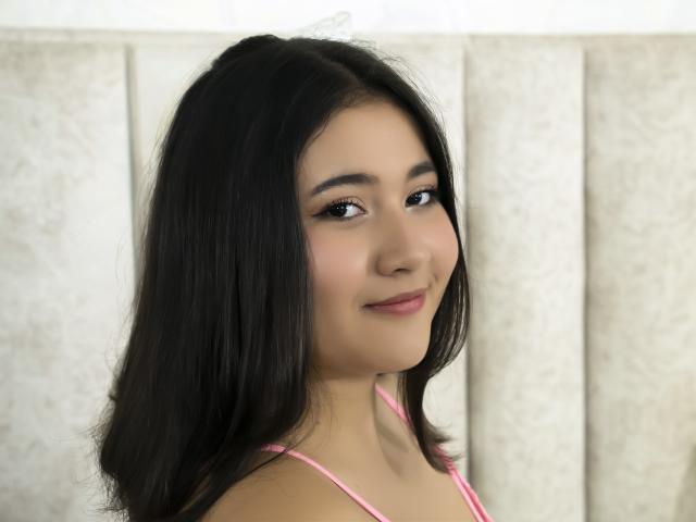CatalinaRosel has dark, straight hair, light brown skin, and a small smile. She's wearing a pink spaghetti strap top, looking over her shoulder with subtle makeup.