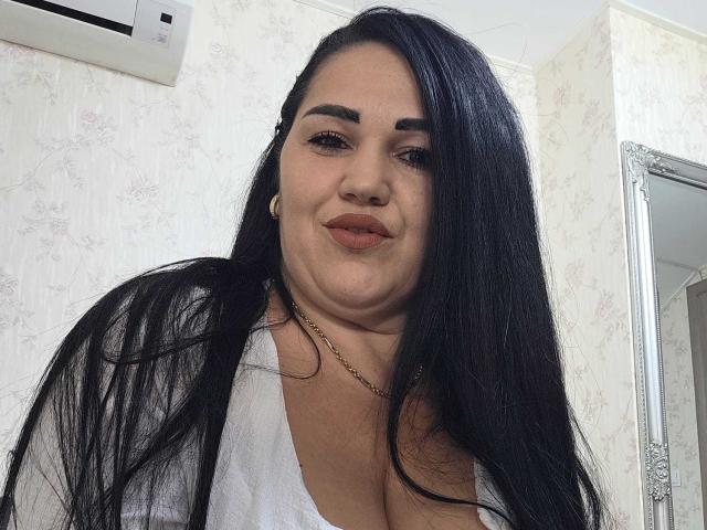 VickyDivinee tem cabelos pretos longos, sobrancelhas grossas e está vestindo uma camisa branca com um decote baixo, que mostra seus seios grandes. Ela está sorrindo levemente, com um rosto bonito..