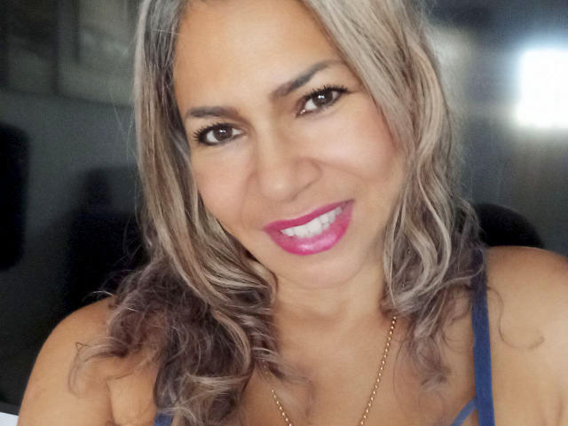 BrigiteWalsh with wavy, gray-brown hair, pink lipstick, and blue top, smiling at the camera. She has brown eyes, dark eyebrows, and a gold necklace.