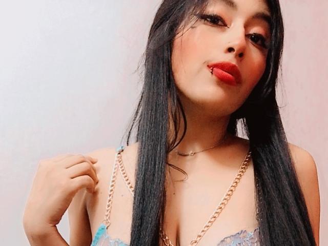 LucianaHernandez with long black hair, red lipstick, and blue lace bra, holding a gold chain, showing off her tits, and giving a pouty expression.