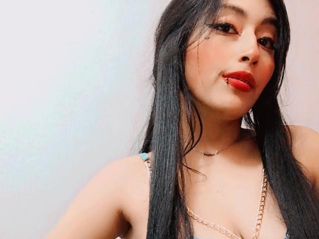 LucianaHernandez with long black hair, red lipstick, and a gold necklace, pouting with a small lip piercing, wearing a low-cut top showing cleavage.