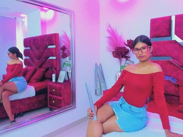 EvaRoux with red off-shoulder top, blue skirt, glasses, holding feather duster, sitting on bed, mirror reflection, pink and purple lighting, red bed, white wall.