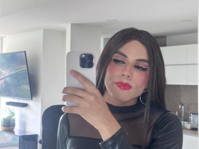 SahraValley with glossy pink lipstick, black leather top, hoop earrings, and pink eyeshadow, taking a selfie with an iPhone in a modern, white kitchen.