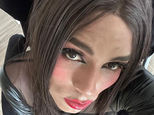 SahraValley has dark brown hair, pink blush, red lipstick, and black eyeliner. She's looking up with green eyes, wearing a black leather top, and has a slight pout.