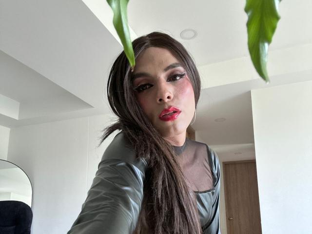 SahraValley with long brown hair, red lipstick, and green leather top, showing off her tits, looking at the camera with a plant in the foreground.