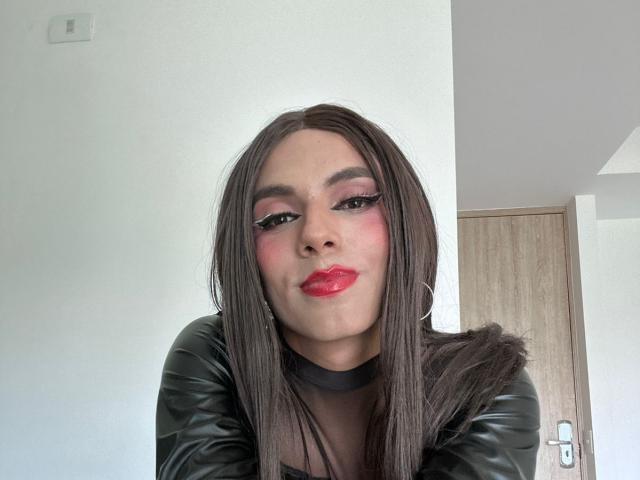 SahraValley, avec de longs cheveux bruns, du rouge à lèvres rouge, du fard à paupières rose, un haut en cuir noir et des boucles d'oreilles cerceaux, souriant légèrement, regardant la caméra, dans une pièce aux murs blancs avec une porte en bois..
