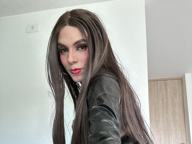 SahraValley with long brown hair, red lipstick, black leather jacket, and pink eye makeup, looking over her shoulder, showing her tits, in a bright room.