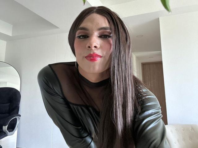 SahraValley with long brown hair, wearing a black sheer leather top, pouting red lips, looking at camera, black eyeliner, white room, round mirror, black chair.