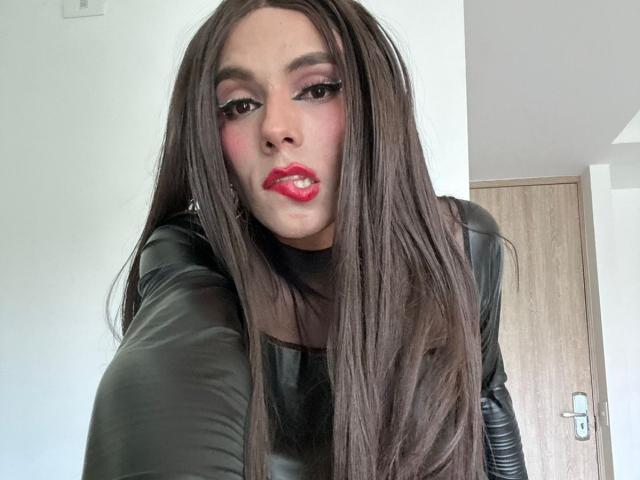 SahraValley, avec de longs cheveux bruns, un haut en cuir noir, du rouge à lèvres et de l'eye-liner, faisant une moue légère, sur une photo de profil. Ses seins sont visibles, mais ne sont pas le point central..