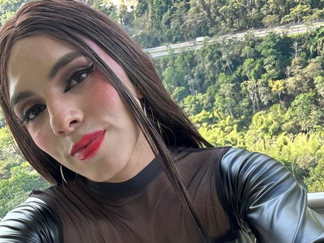 SahraValley has red lipstick, black winged eyeliner, and long brown hair, wearing a sheer black top with black leather sleeves, looking at the camera with a slight smile.