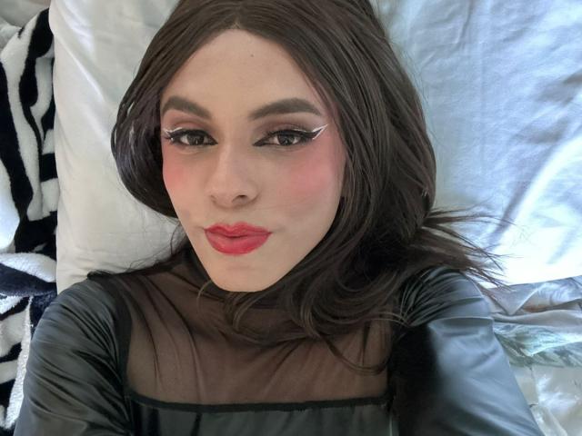 SahraValley has red lipstick, black eyeliner wings, and a cute face with pink cheeks, lying on a bed with white pillows, wearing a black sheer top.
