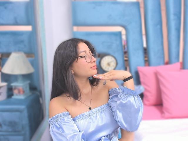 Genesiss with long black hair, glasses, and light blue off-shoulder dress, touching her chin, showing a small tattoo on her right shoulder, sitting in a blue room.