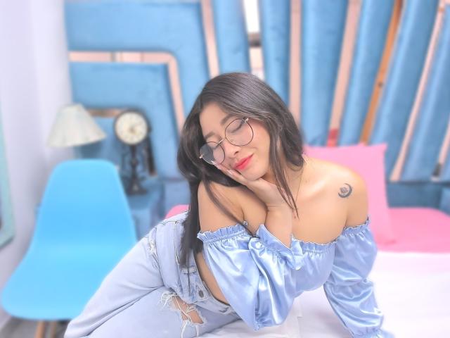 Genesiss with glasses, red lipstick, and blue off-shoulder top, showing a small crescent tattoo on her right shoulder, leaning on a bed with light blue and pink pillows.