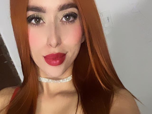 SarahLuxe has red hair, green eyes, and red lipstick. She's wearing a sparkly choker and has a cute smile. Boobs are not visible. She looks sexy and pretty.