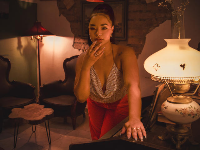 MilaRomano with red pants, silver halter top, and her hand near her mouth, standing next to a lamp, showing her tits, in a warmly lit room.