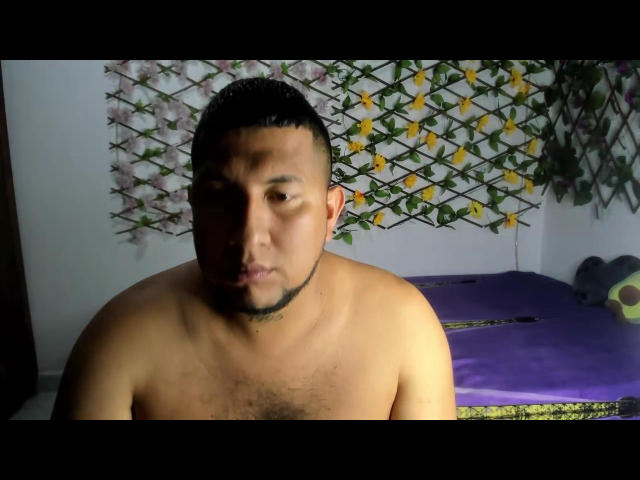 Shirtless ElianGoldS with a light brown skin tone, short black hair, and a beard, making a pouty face, chest hair visible, purple bed with yellow and green patterned wall.