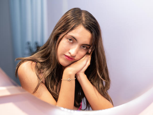 DaniMontolla with long brown hair, looking at camera, hands under chin, subtle smile, pink lipstick, bracelet on right wrist, soft light, pink and white background.