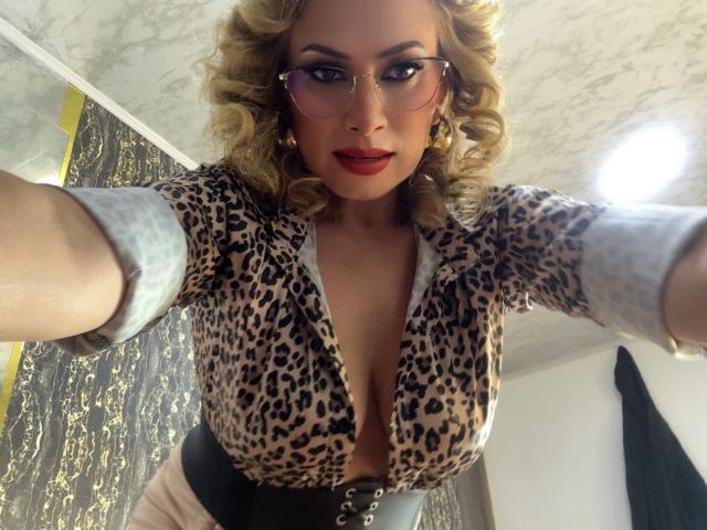MisseScarleth with curly blonde hair, wearing glasses, leopard print top, deep cleavage, black belt, taking a selfie, smiling, red lipstick, marble ceiling, and a ring on her right hand.