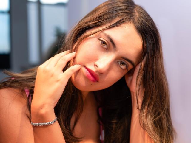 DaniMontolla with brown hair, pink lips, and a silver bracelet, touching her face with a slightly melancholic expression, in a tight, colorful top. Her tits are barely visible.