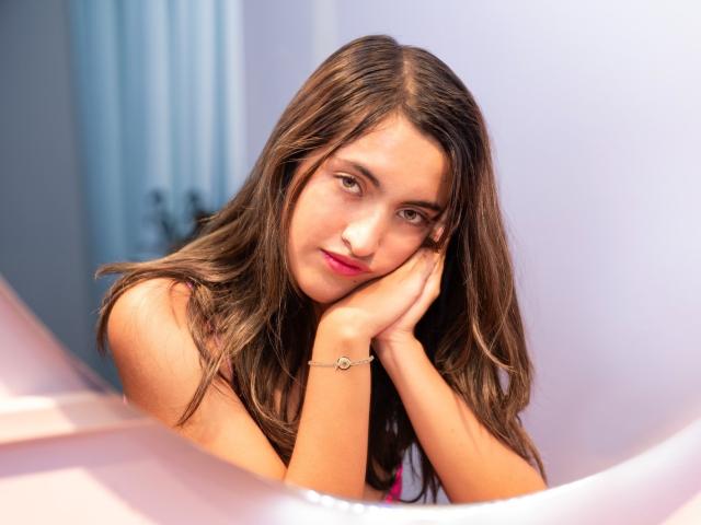 DaniMontolla with long brown hair, pink lips, and a cute smile, resting her head on her hand, wearing a bracelet, in a soft-lit, slightly blurred shot.