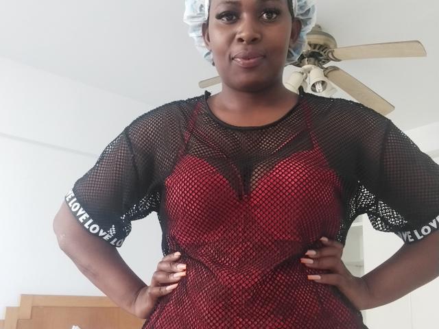 Saltryhips69 está de pie con las manos en las caderas, vistiendo un vestido de malla roja con mangas negras, malla negra sobre un sujetador rojo, y una red para el cabello azul, sonriendo con confianza..