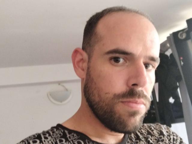 TitiHotMan has a bald head, short brown beard, and is wearing a black and white patterned shirt, looking serious with a slight frown.