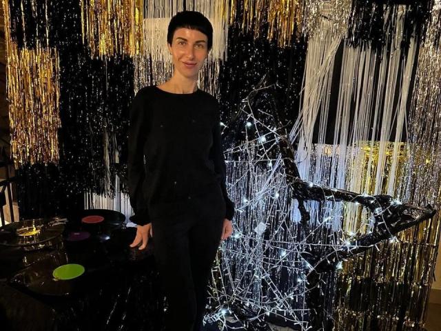 ViolaTray stands with short black hair, wearing a black long-sleeve top and black pants, in front of a shiny, reflective metal sculpture, smiling subtly.
