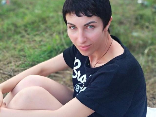 ViolaTray with short black hair, green eyes, and a slight smile, sitting cross-legged in a black t-shirt, looking up with a relaxed, cute expression.