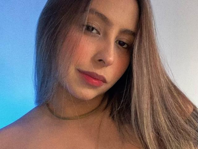 NicoleSweetyPie with long brown hair, pink lips, and a subtle smile, wearing a green choker, looking slightly to the side. Her tits are not visible.