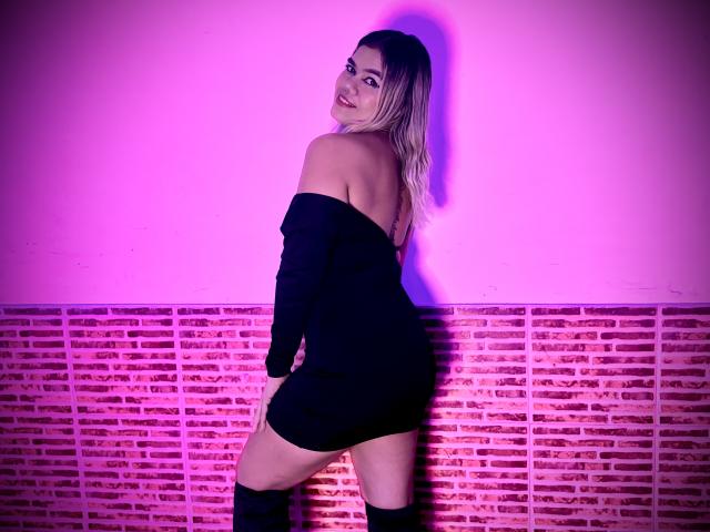 TarahDustin with blonde hair, smiling, wearing an off-shoulder black dress and knee-high boots, standing with her back to the camera, showing her ass. Purple-pink lighting.