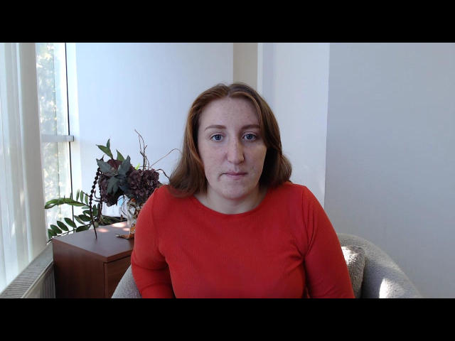 LiliTrayya has red hair, freckles, and is wearing a red long-sleeve shirt, sitting in a gray chair with a wooden table and vase of flowers in the background.