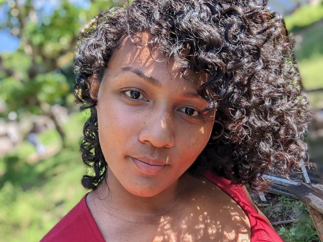 Ninah69s has curly black hair, medium brown skin, and is wearing a red shirt. She's smiling slightly with a sunlit, green, blurred background. Her eyes are focused on camera.