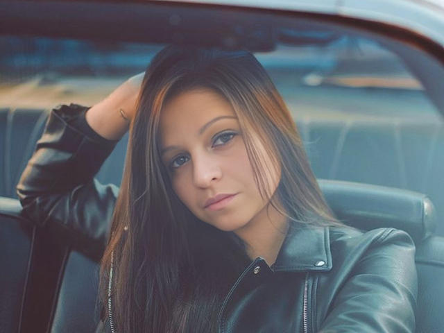 CatMelissa, con lunghi capelli castani, indossa una giacca di pelle nera, ha una mano tra i capelli, appare seria, è seduta in un'auto, ha gli occhi azzurri, l'illuminazione è soffusa, è sexy..