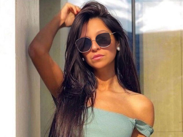 CatMelissa, con cabello largo y oscuro, usando gafas de sol tipo aviador, una blusa verde con un hombro descubierto y aretes de perlas, está de pie con el brazo levantado. Se ven parcialmente los senos. Tiene la piel bronceada, labios rosados y una expresión segura..