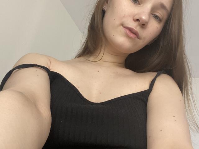 HotCaramells with light skin, brown hair, and small moles, wearing a black ribbed top with thin straps, looking down at the camera with a slight smile.