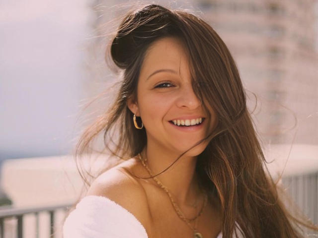 CatMelissa mit langen, braunen Haaren, goldenen Reifenohrringen, die mit weißen Zähnen lächelt, trägt ein weißes Oberteil mit einem Schulterauschnitt und eine goldene Halskette und hat ein süßes, attraktives Gesicht..