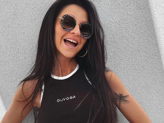 CatMelissa with long black hair, smiling with mouth open, wearing round sunglasses, black sleeveless top, and hoop earrings, has a tattoo on her right arm, and is tan.