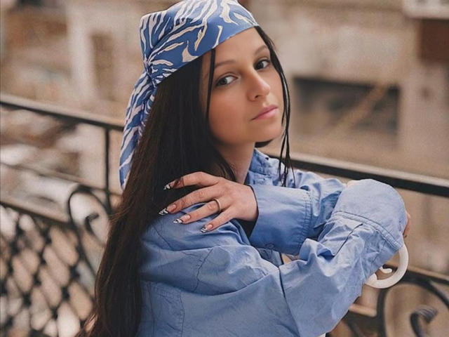 CatMelissa with long dark hair, blue patterned headscarf, and blue jacket, arms crossed, looking over shoulder, nails with white and silver designs, subtle makeup, outdoor setting.