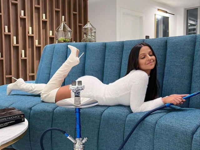 CatMelissa, con lunghi capelli neri, indossa un abito bianco e vaporoso e stivali bianchi che arrivano alle cosce, ed è sdraiata su un divano blu, fumando una shisha con un tubo blu..