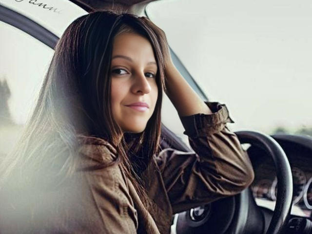 CatMelissa with long brown hair, wearing a brown jacket, sits in a car, hand in hair, smiling slightly, looking to the side, with a relaxed, confident expression.