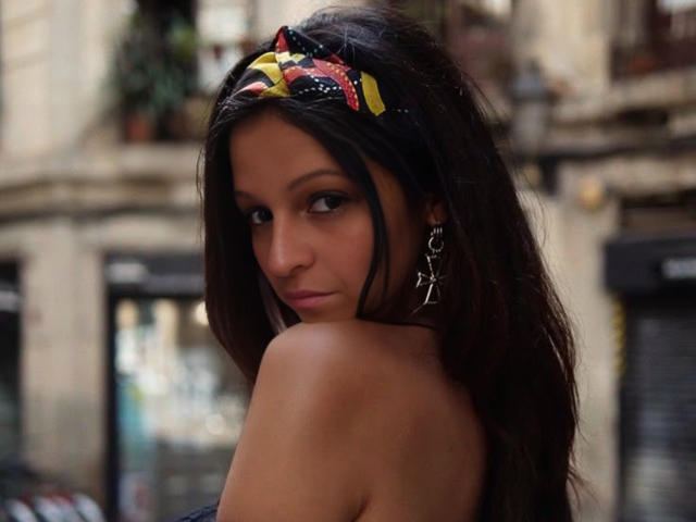 CatMelissa with long dark hair, wearing a colorful headband and star earrings, looking back with a slight smile, showing her bare shoulder and small tits.