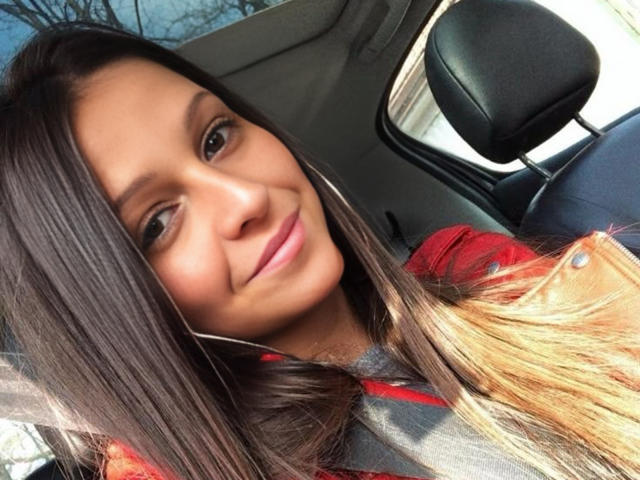 CatMelissa with long brown hair, light brown eyes, and a small smile, wearing a red shirt, seated in a car with a black headrest in the background.
