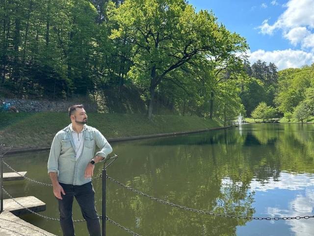 NormanGlory, standing by a calm river, wearing a light blue jacket and black pants, with a chain fence in the foreground, and lush green trees in the background.