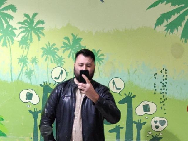 NormanGlory with a black beard and leather jacket, holding a phone to his mouth, standing in front of a green and yellow wall with musical notes and symbols.